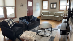 Una sala de estar con un sofá negro, un sofá seccional color beige, una pequeña mesa redonda y una silla de madera.