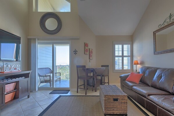 A living room with a brown leather couch, a wicker ottoman, a dining table with chairs, a TV on a stand, and a large mirror on the wall. - 423 Grand Caribbean Beach Condo - Amazing location with access to everything (Orange Beach)