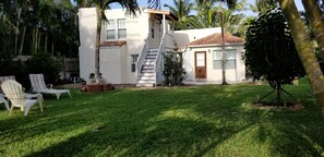 A two-story house with a white exterior, a red-tiled roof, and a staircase leading to the upper floor. There are white lounge chairs and a small garden area with a palm tree.