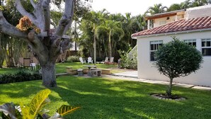Un jardin bien entretenu, avec un espace patio, un petit arbre et un bâtiment blanc surmonté d’un toit recouvert de tuiles rouges.