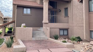 A multi-story residential building with a brick facade, a balcony with a metal railing, and a small garden area with rocks and plants. - Sparkling Newly Remod Condo. Near Zion's, Tuacahn, Bike Trails. Pool &Pickleball (St George)