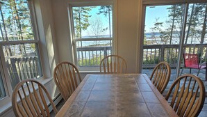 A dining area with a table and chairs, large windows with a view of trees and a body of water, and a wooden deck outside. - Large Oceanfront house in Gouldsboro, Maine near Acadia NP; Views! (Gouldsboro)