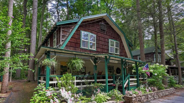 Una casa de madera rústica con un porche, rodeada de vegetación exuberante y un muro de piedra.