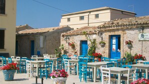Un espace extérieur aménagé avec des tables et des chaises, un bâtiment en pierre avec une porte bleue, et des plantes en pot ornées de fleurs rouges.