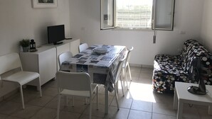 Un coin repas avec une table recouverte d’un journal, des chaises blanches, un téléviseur posé sur un meuble et un canapé à motifs à pois.