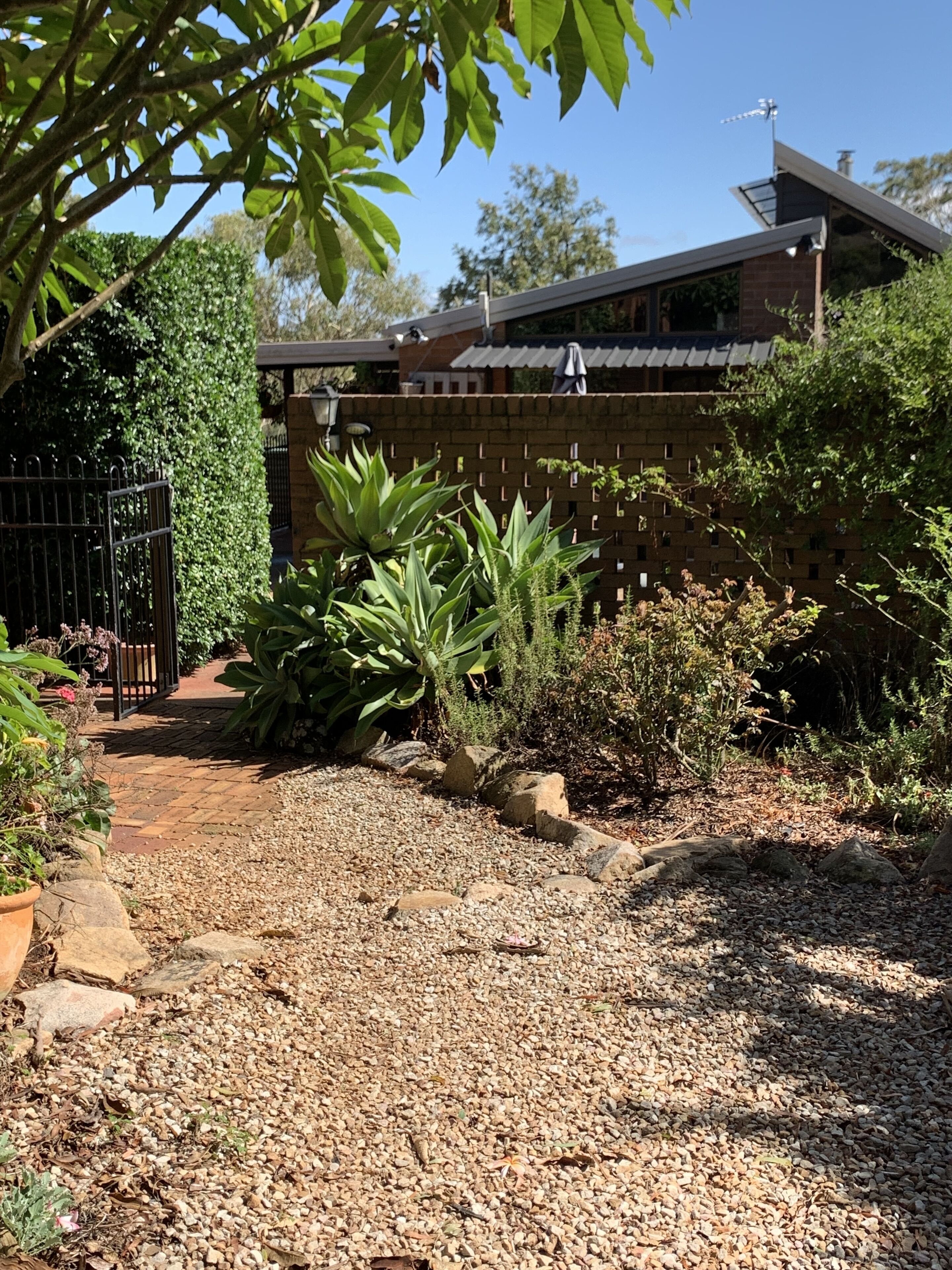 Un jardin avec un chemin de gravier, différentes plantes, un mur de briques et une maison avec un toit en pente.