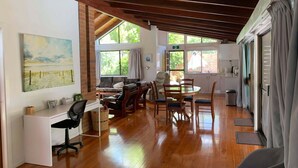 A spacious room with a wooden ceiling, a dining area with a round table and chairs, a sofa, a desk with a computer, and large windows providing natural light. - Cranley Garden Retreat with Fireplace & Pool (Cranley)