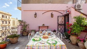 Una terraza en la azotea con una mesa dispuesta para una comida, rodeada de plantas en macetas y un muro rosa.