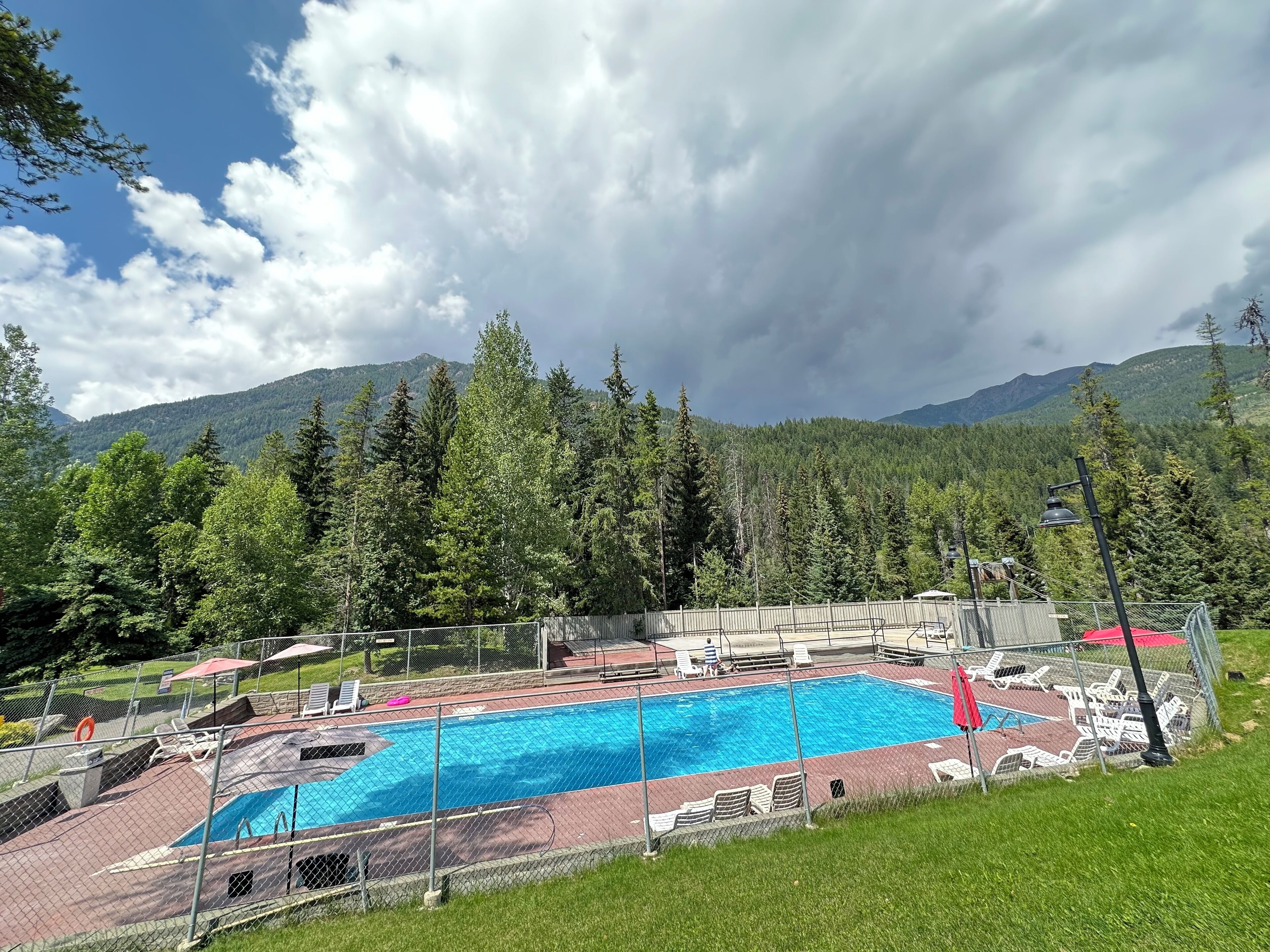A swimming pool with a fenced deck, surrounded by trees and mountains.