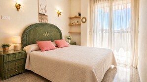 Une chambre à coucher comprenant un lit, des tables de chevet, une lampe, une horloge murale, une fenêtre avec des rideaux légers et une étagère fixée au mur, ornée d’objets décoratifs.