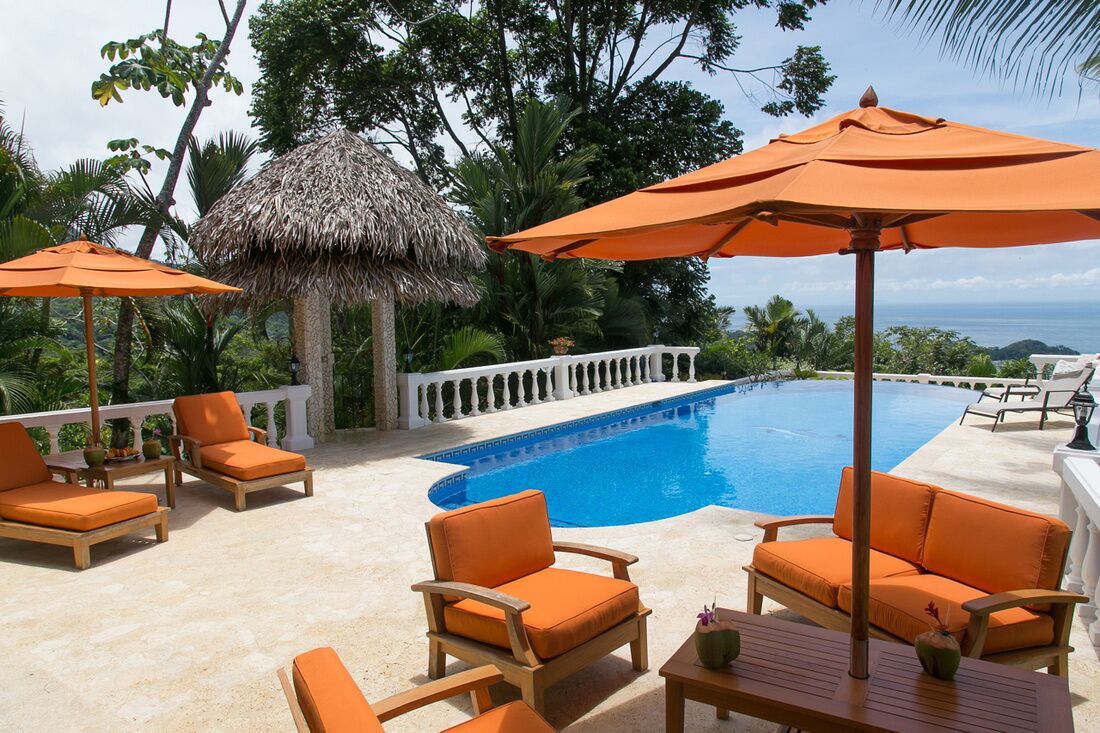 Un espace aménagé au bord de la piscine, avec des chaises et des parasols orange, une structure recouverte de chaume et une vue sur l’océan.