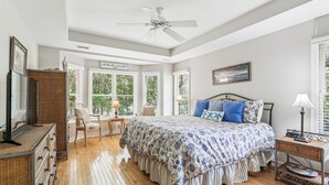 A bedroom with a large bed, a ceiling fan, a desk with a chair, and a view of greenery outside.