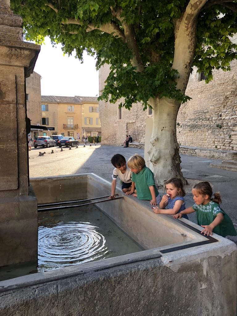 Un groupe d’enfants joue près d’une fontaine dans une cour extérieure, avec des bâtiments en pierre en arrière-plan.