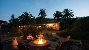 A fire pit with chairs around it, a house in the background, and palm trees.