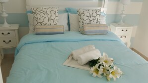 A neatly made bed with a light blue bedspread, two pillows with a floral pattern, a folded white towel, and a bouquet of white flowers.