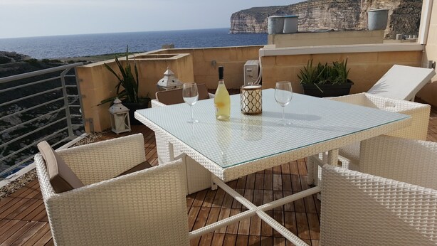 A white wicker table with glass top, surrounded by white wicker chairs, on a wooden deck overlooking the sea.