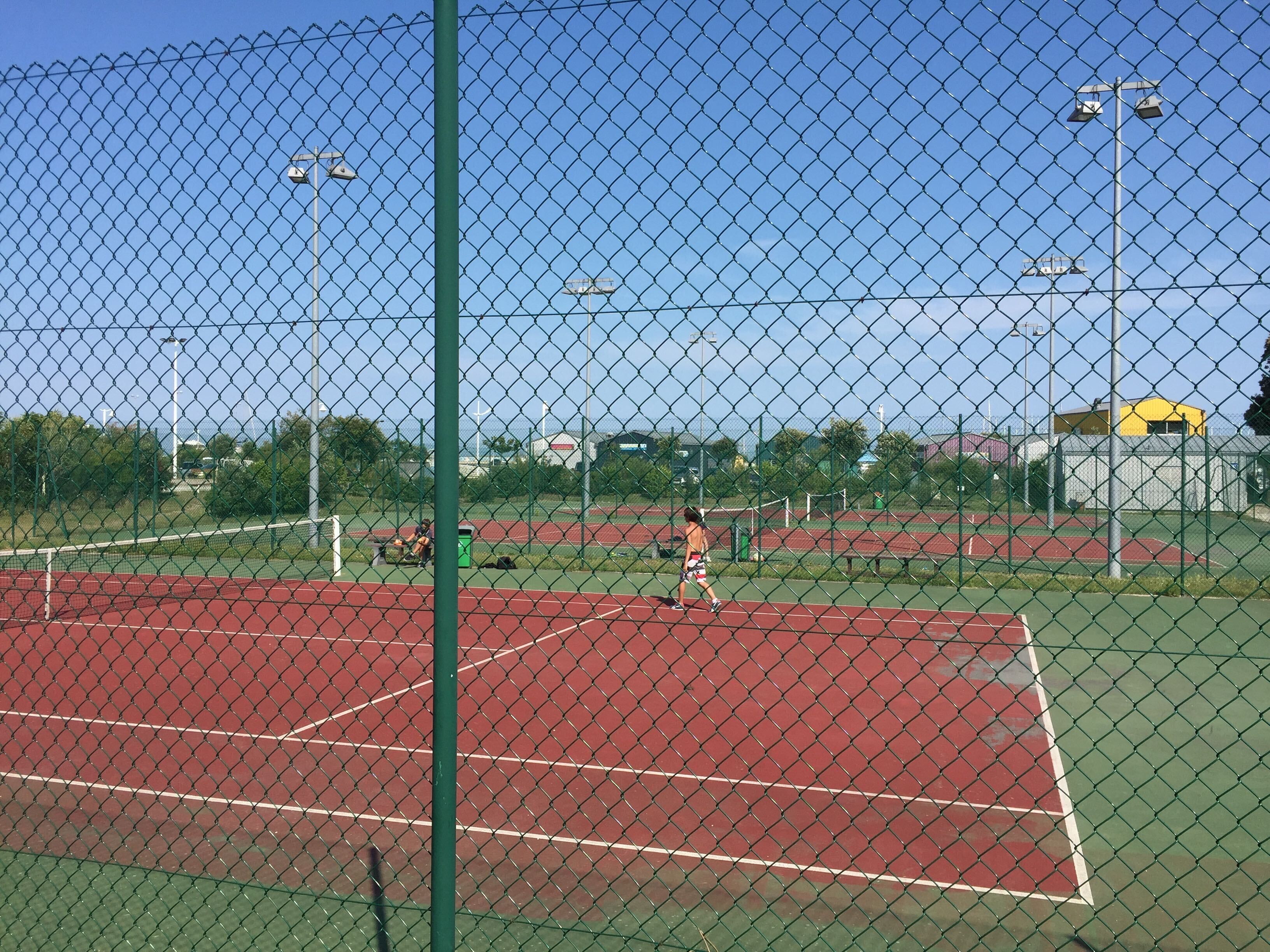 A tennis court with a chain-link fence, multiple tennis nets, and floodlights.
