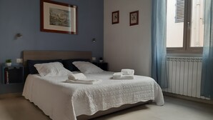 Una cama bien hecha con edredón blanco y almohadas, una mesita de noche con una planta y un cuadro en la pared.
