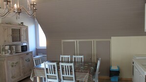 A dining area with a table set for four, surrounded by white chairs. A chandelier hangs above the table, and a microwave is visible on a cabinet to the left.