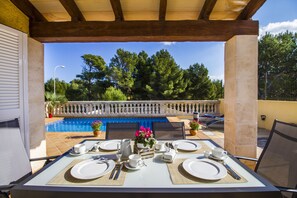 An outdoor dining area with a view of a swimming pool, surrounded by greenery and a wooden pergola. - Villa del Mar / Beach house (Capdepera)