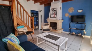 Un salon comprenant un canapé, deux fauteuils en osier, une table basse blanche, une cheminée, un téléviseur et une bibliothèque.