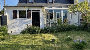 Une maison à deux étages, avec une façade blanche, un toit foncé et un petit jardin agrémenté de pelouse et de fleurs.