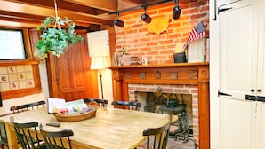 Un comedor rústico con una mesa y sillas de madera, una planta colgante, una chimenea de ladrillo y una estantería con libros y una bandera estadounidense.