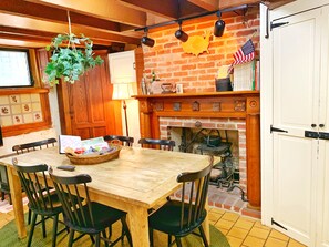 A rustic dining area with a wooden table and chairs, a hanging plant, a brick fireplace, and a bookshelf with books and an American flag.
