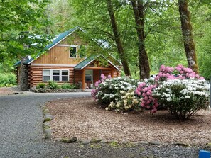 Une cabane en rondins avec une cheminée en pierre, entourée d’une végétation luxuriante et de buissons en fleurs.