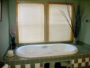 Une salle de bain avec une grande baignoire, deux fenêtres équipées de stores, une plante en pot et un sol carrelé.