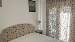 Une chambre à coucher avec un lit, une table de chevet, une lampe, un tableau encadré au mur et un rideau orné d’un motif marin.