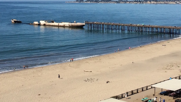 Une plage avec une jetée qui s’avance dans l’eau, un rivage sablonneux et une ligne côtière lointaine parsemée de bâtiments.