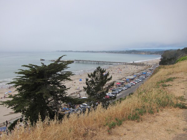A coastal beach with a long pier extending into the water, a parking lot with cars, and a grassy hill in the foreground.