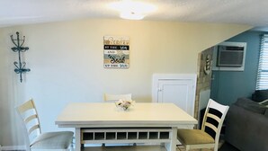 A dining area with a white table, two chairs, a decorative wall piece, and a grey couch in the background. - A Stone Throw Away from the Beach (Wildwood)