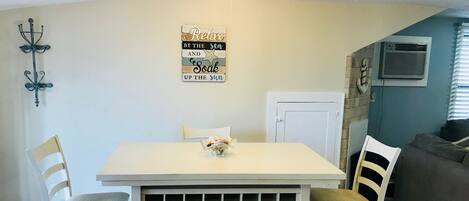 A dining area with a white table, two chairs, a decorative wall piece, and a window with blinds.