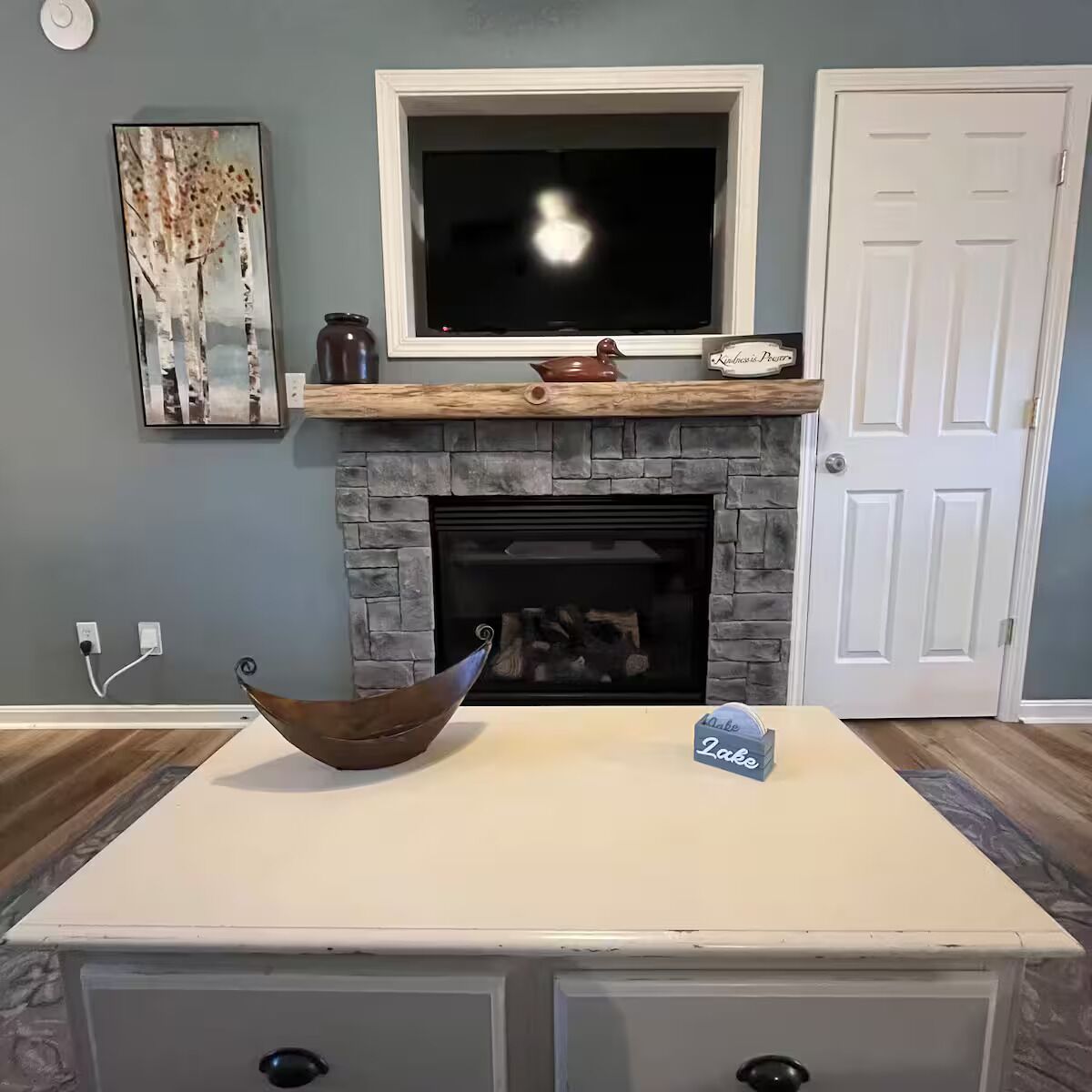 Una sala de estar con chimenea, un televisor encima, una puerta a la derecha y una mesa blanca con un cuenco y una caja.