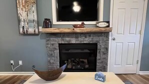A living room with a fireplace, a television above it, a door to the right, and a white table with a bowl and a box on it. - Lakeside Haven, Cottage#4, Pool, Hot Tub, Lake View (Taylorsville)