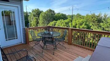 A wooden deck with a glass table and chairs, a barbecue grill, and a view of trees.