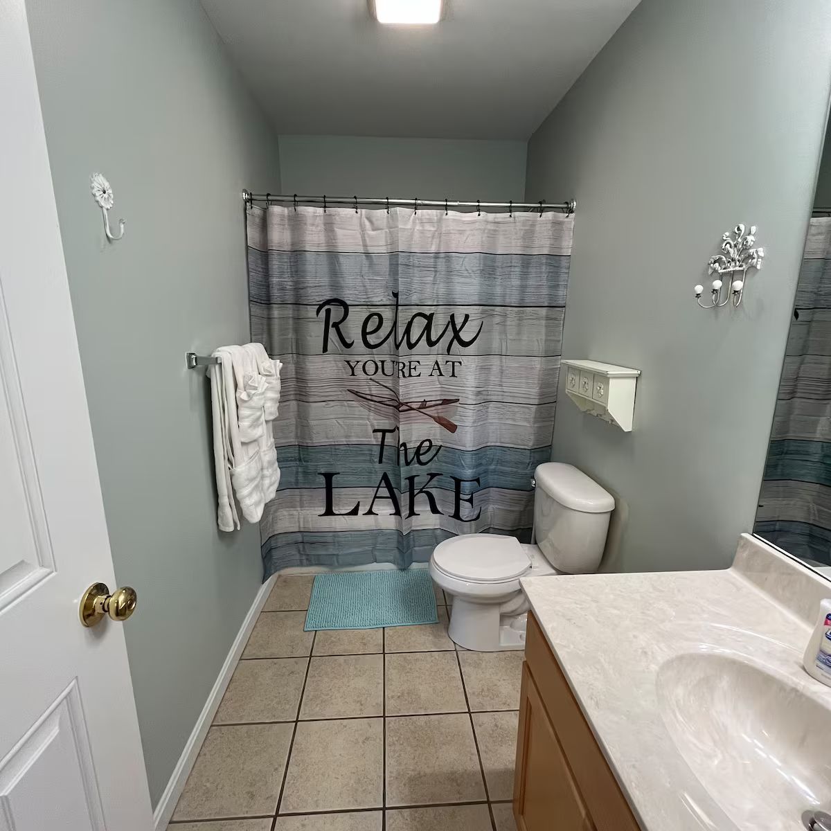 Une salle de bain avec un rideau de douche sur lequel est écrit « Détendez-vous, vous êtes au bord du lac », des toilettes, un lavabo avec un miroir et un porte-serviettes.