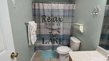 Une salle de bain avec un rideau de douche sur lequel est écrit « Détendez-vous, vous êtes au bord du lac », des toilettes, un lavabo avec un miroir et un porte-serviettes.