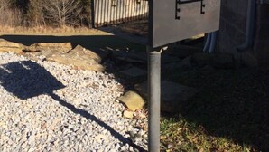 A metal mailbox attached to a metal pole, with a wooden bench in front of a house.
