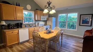 A kitchen with wooden cabinets, a dining table with chairs, a chandelier, and a view of greenery outside.