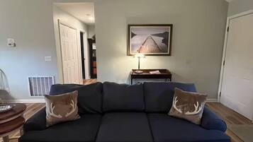 A living room with a dark blue sofa, two decorative pillows, a framed picture on the wall, and a small table with a lamp.