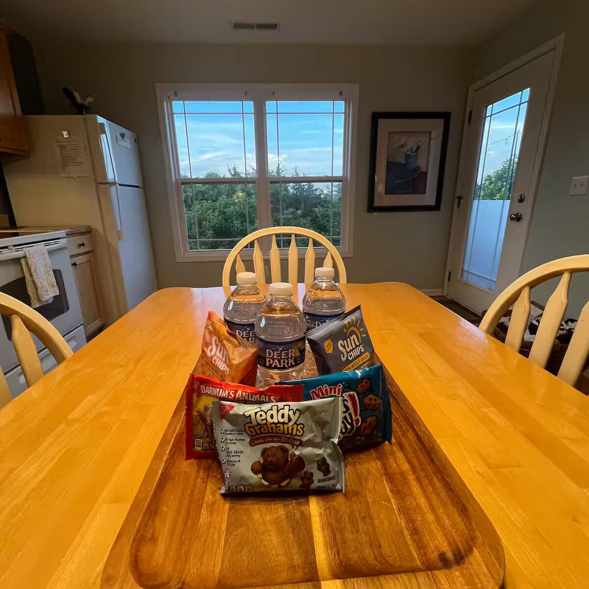 A wooden dining table with a tray holding snacks and drinks, a refrigerator, and a window with a view of trees.