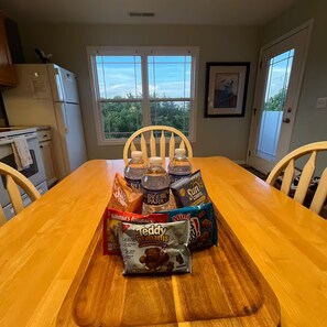 A wooden dining table with a tray holding snacks and drinks, a refrigerator, and a window with a view of trees.