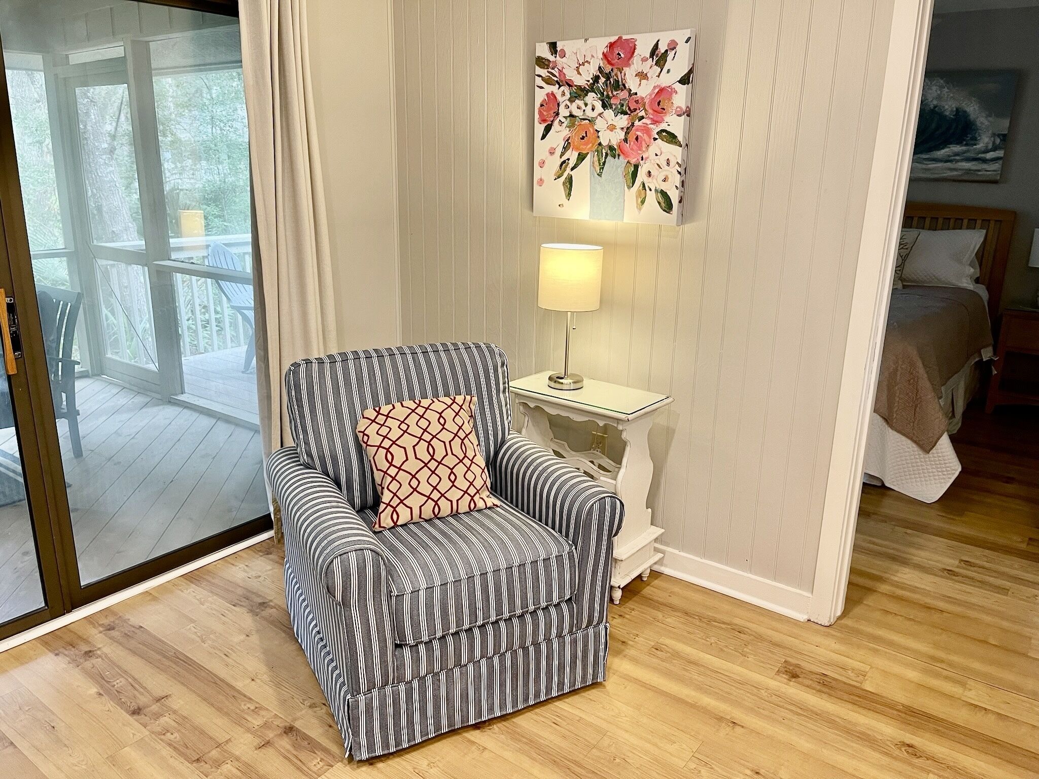 Un sillón a rayas con un cojín estampado, una mesita de noche con una lámpara, y un cuadro floral en la pared.