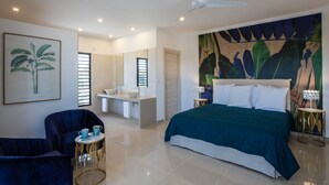Une chambre moderne dotée d’un grand lit, d’un coin salon avec un fauteuil et une table, d’une salle de bain avec un lavabo et un miroir, et d’un ventilateur de plafond.