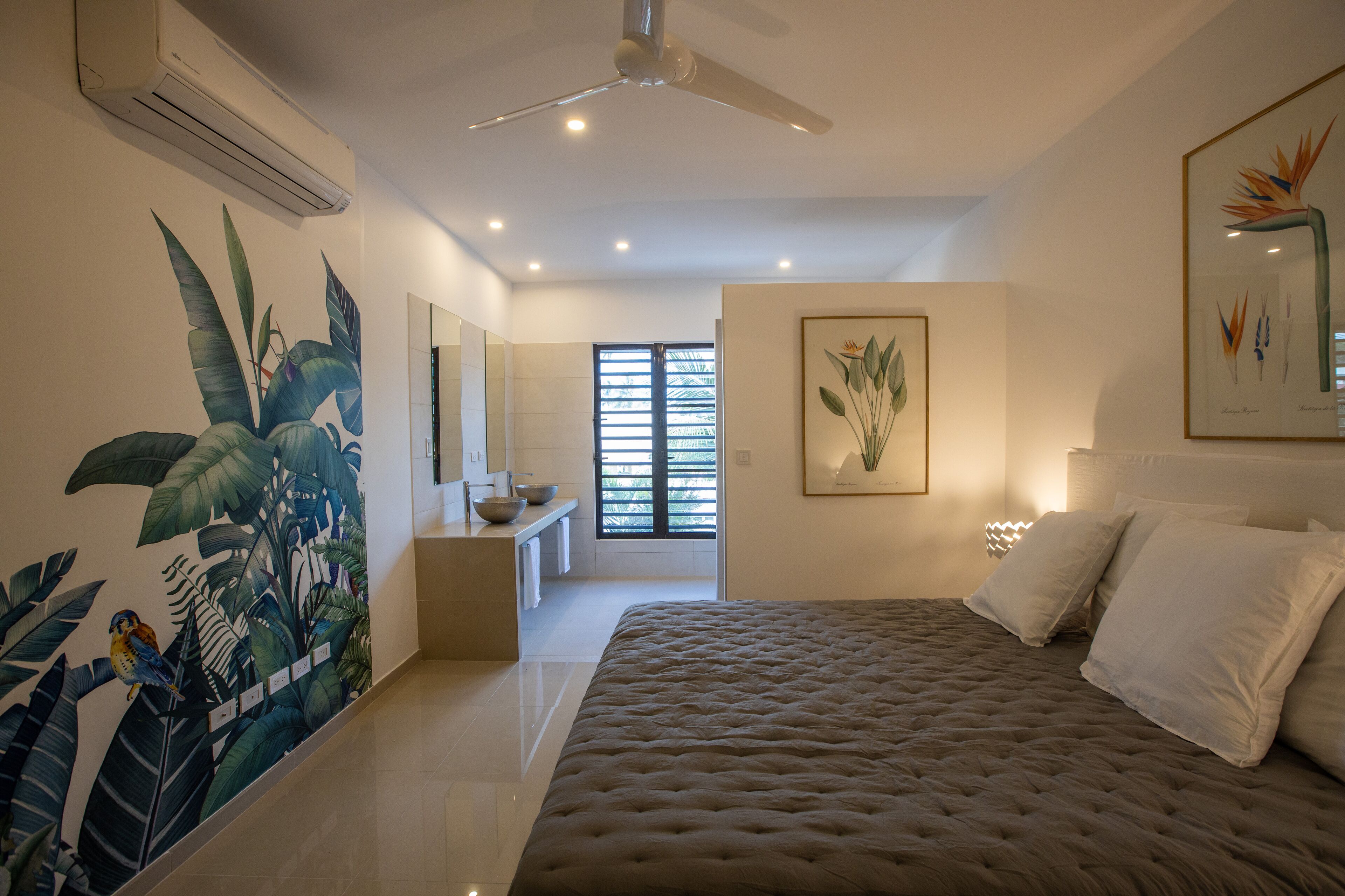 A bedroom with a large bed, a ceiling fan, a wall-mounted air conditioner, a window with blinds, and a bathroom with a sink and mirror.