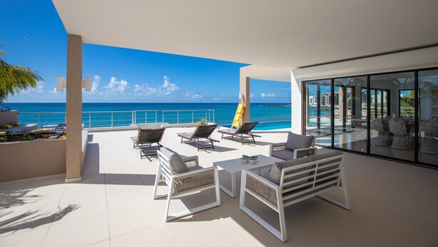 Une vaste terrasse extérieure offrant une vue sur l’océan, aménagée avec des chaises et des tables blanches, et une porte vitrée donnant accès à un espace intérieur.