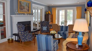 Een woonkamer met een open haard, blauw-wit gestreepte fauteuils, een blauwe lamp en een schilderij aan de muur.